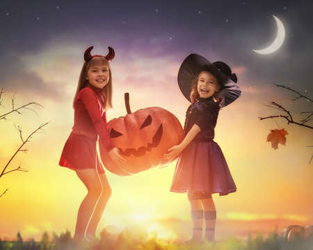 Two happy sisters on Halloween. Funny kids in carnival costumes outdoors. Cheerful children and pumpkin on sunset background.の写真素材