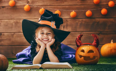 Happy Halloween! Cute cheerful little witch with a book of spells. Beautiful child girl in witch costume sitting on grass, reading and laughing.の写真素材