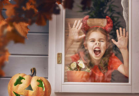 Happy Halloween! Cute cheerful kid looking on the window. Beautiful child girl in devil costume in the house decorated to Halloween.の写真素材