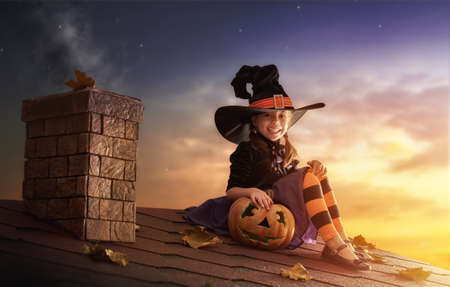 Happy Halloween! Cute little witch with a pumpkin. Beautiful young child girl in witch costume sitting on the roof.の写真素材