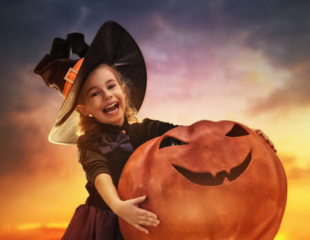 Happy Halloween! Cute little witch with a big pumpkin. Beautiful young child girl in witch costume outdoors.の写真素材