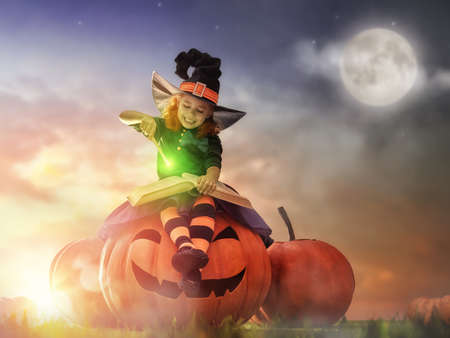 Happy Halloween! Cute cheerful little witch with a magic wand and book of spells. Beautiful child girl in witch costume sitting on the big pumpkin, conjuring and laughing.の写真素材