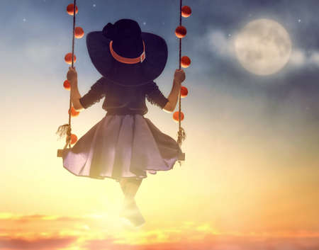 Happy Halloween! Cute little witch on swing in sunset. Beautiful young child girl in witch costume outdoors.の写真素材