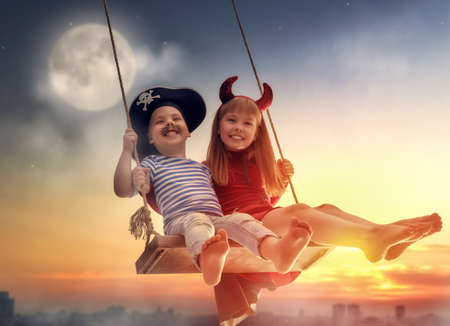 Two happy children on Halloween. Funny kids in carnival costumes outdoors. Cheerful boy and girl on swings on sunset background.の写真素材