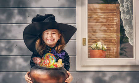 Happy Halloween! Cute cheerful little witch outdoors. Beautiful child girl in witch costume with candy near house.の写真素材