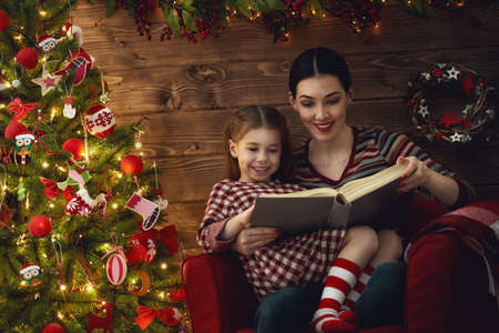 Merry Christmas! Pretty young mother reading a book to her daughter near Christmas tree.の写真素材