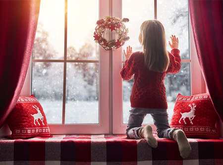 Merry Christmas and happy holidays! Cute little girl sitting by the window and looking at the winter...