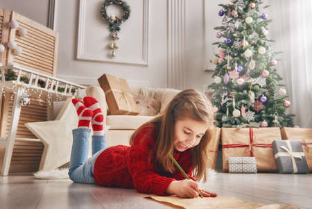 Merry Christmas and Happy Holidays! Cute little child girl writes the letter to Santa Claus near Christmas tree indoors.の写真素材