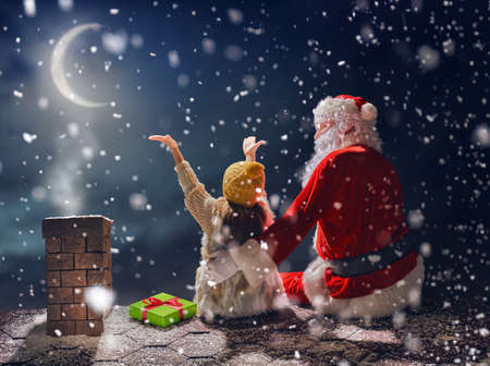 Merry Christmas and happy holidays! Cute little child girl and Santa Claus sitting on the roof and looking at snowfall. Christmas legend concept.の写真素材