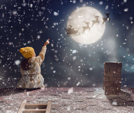 Merry Christmas and happy holidays! Cute little child girl sitting on the roof and looking at Santa Claus flying in his sleigh against moon sky. Kid enjoy the holiday. Christmas legend concept.の写真素材