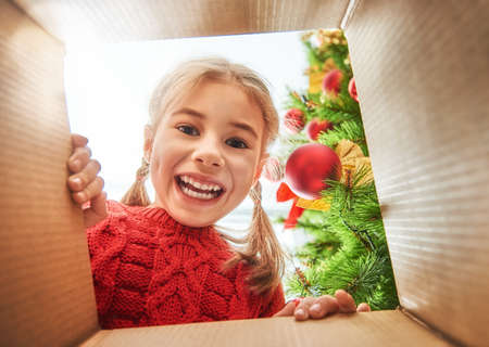 Merry Christmas and Happy Holidays! Cheerful cute child girl opening a Christmas present. Little kid having fun near Christmas tree indoors. View from inside of the box.の写真素材