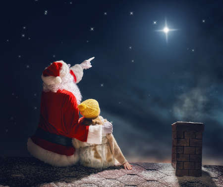 Merry Christmas and happy holidays! Cute little child girl and Santa Claus sitting on the roof and looking at Christmas star. Christmas legend concept.の写真素材