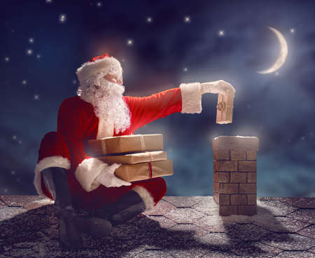 Merry Christmas and happy holidays! Santa Claus sitting on the roof of the house and puts the presents in the chimney. Christmas legend concept.の写真素材