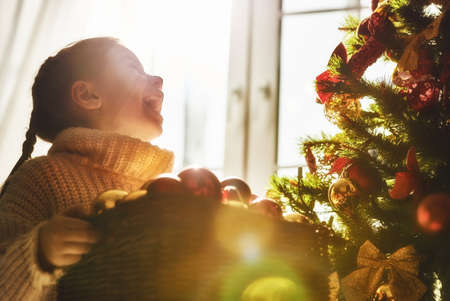 Merry Christmas and Happy Holidays! Cute little child girl is decorating the Christmas tree indoors. The morning before Xmas. Portrait kid close up.の写真素材