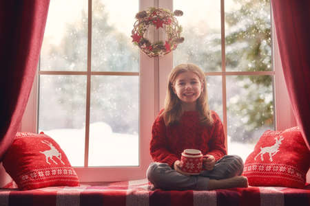 Merry Christmas and happy holidays! Cute little girl sitting by the window with a cup of hot drink and looking at the winter forest. Room decorated on Christmas. Kid enjoys the snowfall.の写真素材