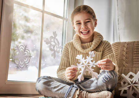 Cute little girl sitting by the window and looking at the winter forest. Child makes paper snowflakes for decoration windows. Kid enjoys the snowfall.の写真素材