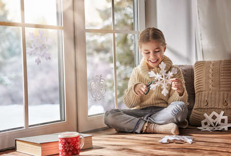 Cute little girl sitting by the window and looking at the winter forest. Child makes paper snowflakes for decoration windows. Kid enjoys the snowfall.の写真素材