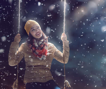 Beautiful young woman on a swing on snowy winter walk. Outdoor fun for winter vacation. Portrait girl on dark background.の写真素材