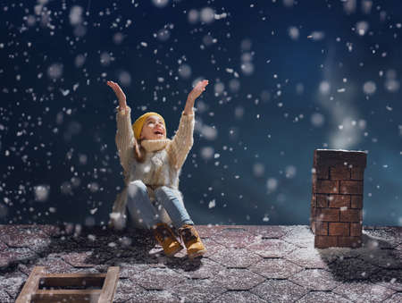 Happy child girl playing on a snowy winter walk. Little girl enjoys the game. Child girl sitting on the roof and looking at snowfall. Outdoor fun for winter vacation. Portrait kid on dark background.の写真素材