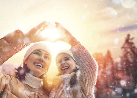 Happy loving family! Mother and child girl having fun, playing and laughing on snowy winter walk in nature. Frost winter season. Mom and kid making hand heart in evening.の写真素材