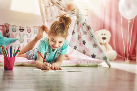 Happy child plays. Little child girl draws with colored pencils indoors. Funny lovely child having fun in children room.の写真素材