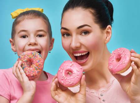 Funny family on a background of bright blue wall. Mother and her daughter girl are having fun with colorful donuts. Dieting concept and junk food. Yellow, pink and turquoise colors.の写真素材