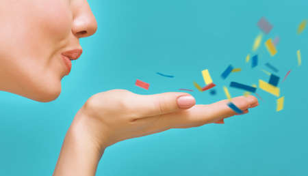 Young woman on background of bright blue wall. Girl blowing multicolor confetti. Close up.の写真素材