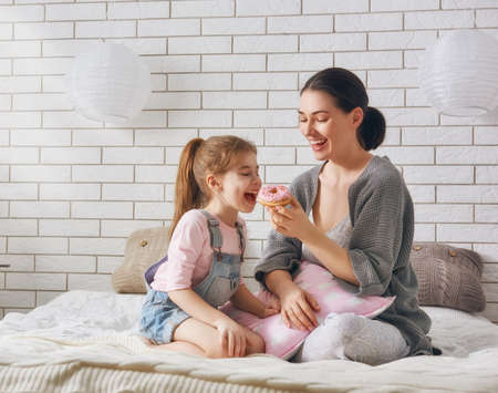 Happy loving family. Mother and her daughter child girl are eating donuts and having fun on the bed in the room.の写真素材