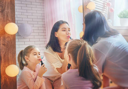 Funny family at home. Mother and her child girl are doing your makeup and having fun near mirror. Mom and daughter wear lipstick in kids room.の写真素材