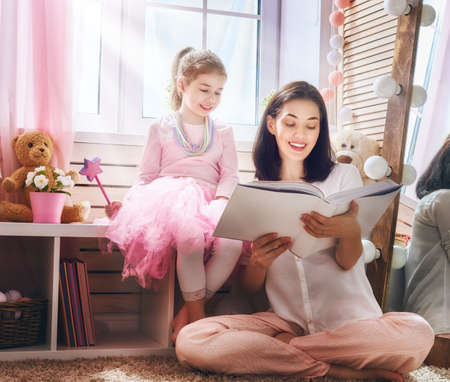 Happy loving family. Pretty young mother reading a book to her daughter indoors. Funny mom and lovely child having fun in children room.の写真素材