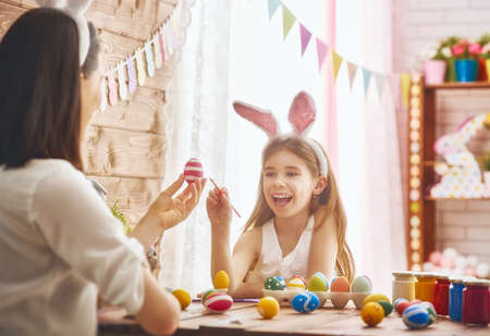 Mother and her daughter painting eggs. Happy family preparing for Easter. Cute little child girl wearing bunny ears.の写真素材