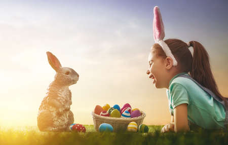 Cute little child wearing bunny ears. Girl has a basket with eggs. Kid sitting on the lawn at Easter in the rays of the setting sun.の写真素材