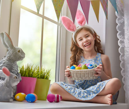 Cute little child wearing bunny ears. Girl sitting on the window with painting eggs.