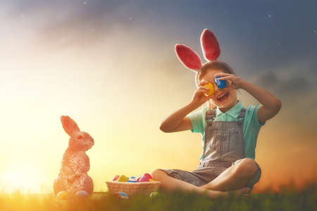 Cute little child wearing bunny ears. Girl has a basket with eggs. Kid sitting on the lawn at Easter in the rays of the setting sun.の写真素材