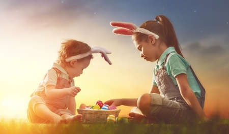 Two cute little children wearing bunny ears. Girls are sitting on the lawn. Kids with Easter eggs in...
