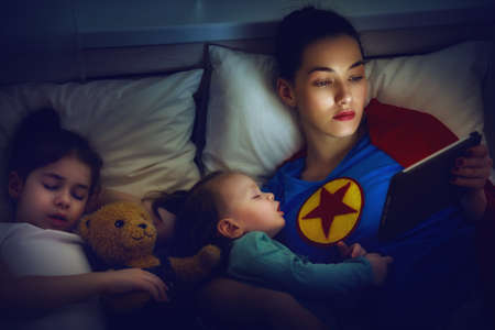 Adorable little children girls are napping in the bed with their mom. Quiet sleep with teddy bear under the protection of the mother superhero.の写真素材
