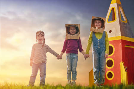 Children in astronauts costumes with toy rocket playing and dreaming of becoming a spacemen. Portrait of funny kids on a background of sunset star sky on nature. Family friends games outdoors.の写真素材