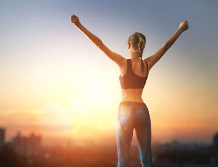 Healthy lifestyle and sport concepts. Woman in fashionable sportswear is doing exercise on nature. Girl with raised hands on sunset background.の写真素材