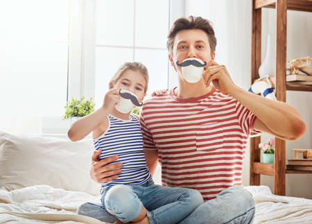 Happy father's day! Dad and his child daughter are playing and having fun together. Beautiful funny girl and daddy have mustaches on cups. Family holidays and togetherness.の写真素材