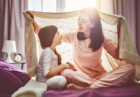A nice girl and her mother enjoy sunny morning. Good time at home. Child wakes up from sleep. Family playing under blanket on the bed in the bedroom.の写真素材