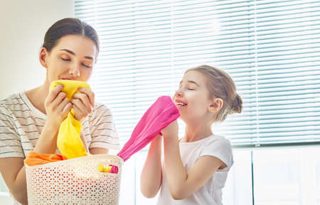 Beautiful young woman and child girl little helper are smelling clean clothes and smiling while doing laundry at home.の写真素材