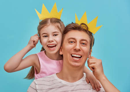 Funny family on a background of bright blue wall. Father and his daughter girl with a paper accessories. Dad and child are holding paper crown on stick.の写真素材