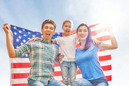 Patriotic holiday. Happy family, parents and daughter child girl with American flag outdoors on blue sky background. USA celebrate 4th of July.の写真素材