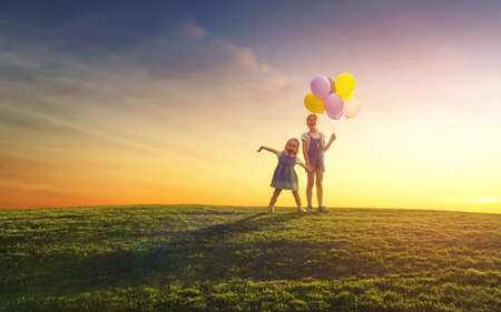 Two happy little children are playing with balloons on meadow at sunset background. Sisters are having fun and laughing on nature in the summer.の写真素材
