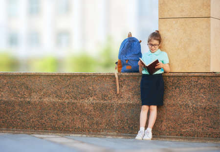 Pupil of primary school with book in hand. Girl with backpack near building outdoors. Beginning of lessons. First day of fall.の写真素材