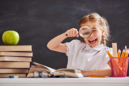Back to school! Happy cute industrious child is sitting at a desk indoors. Kid is learning in class on background of blackboard.の写真素材