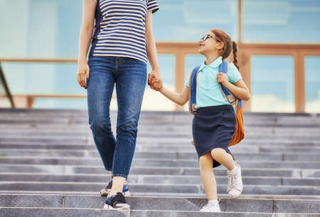 Parent and pupil of primary school go hand in hand. Woman and girl with backpack behind the back. Beginning of lessons. First day of fall.の写真素材