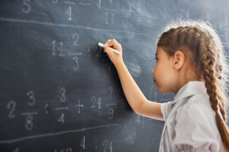 Tough day at school! Cute child near the blackboard indoors. Kid is learning in class. Complex math, arithmetic and examples. Numbers written with chalk on board.の写真素材