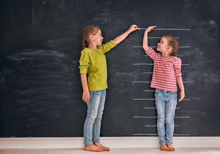 Two children sisters play together. Kid measures the growth on the background of blackboard. Concept of education.の写真素材