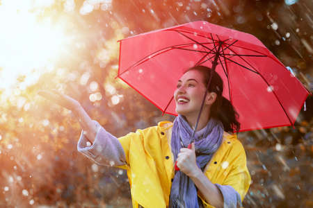 Happy beautiful young woman with red umbrella under the autumn shower. Girl is wearing yellow raincoat and enjoying rainfall. の写真素材